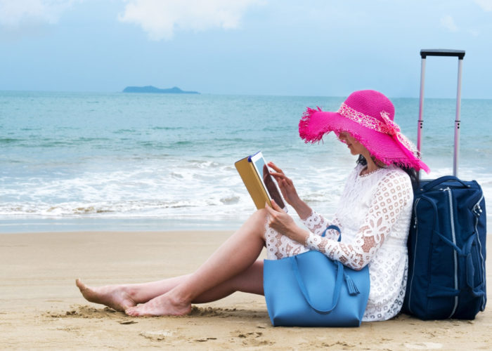 Tourist reclining against her luggage on the beach, reading on her iPad