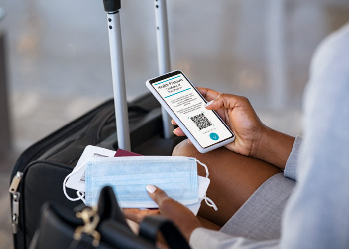 Woman sitting by her luggage, holding a facemask and a phone showing a COVID-19 Health Passport