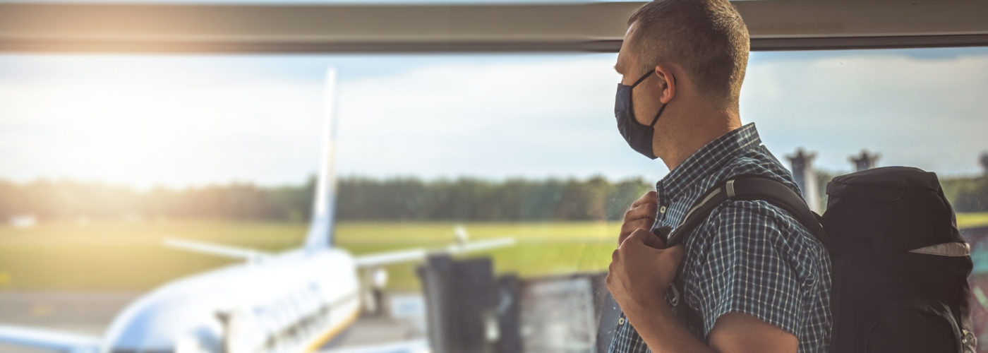 Man wearing a face mask and looking out an airport window at a plane on the tarmac