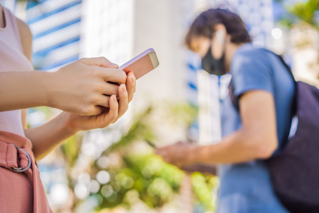 A woman, in focus in the foreground, using her smartphone and a man, out of focus in the background, also using his smartphone and wearing a face mask
