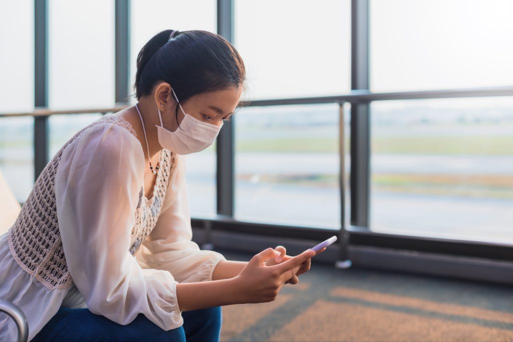 Woman sitting in airport terminal, wearing a face mask and using her smartphone